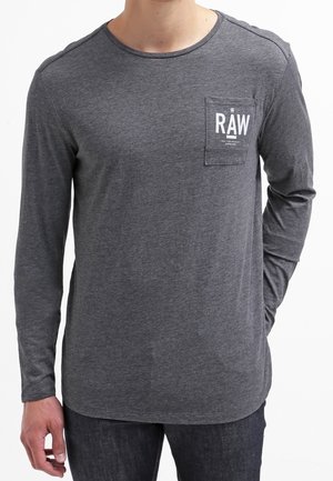 Homme portant une chemise à manches longues gris foncé avec une petite poche poitrine affichant le texte blanc "RAW" sur la poche.
