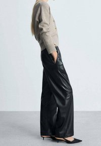 Cardigan beige en tricot avec des boutons dorés, associé à un pantalon large en cuir noir et des escarpins noirs à bout pointu. Vue de profil latéral.