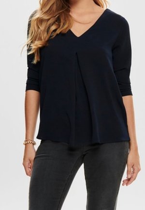 Kvinde iført en marineblå V-hals langærmet top med frontplisse detaljer og mørkegrå tætsiddende jeans, stående med armene afslappet.