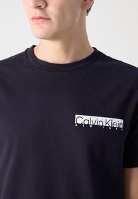 Juodas medvilninis marškinėliai su apvaliu kaklu, su balta grafine logotipu kairėje krūtinės pusėje. Lygus paviršius, trumpomis rankovėmis, kasdieniškas dizainas.