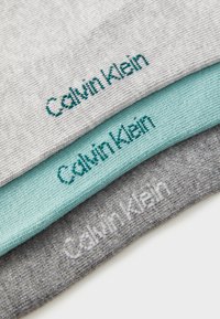 Tres pares de calcetines plegados de Calvin Klein en gris claro, azul verdoso claro y gris oscuro, con logotipos de la marca bordados.