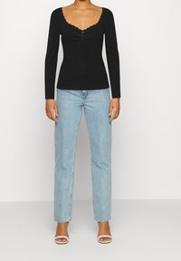 Schwarzes, geripptes Langarmshirt mit wellenförmigem Ausschnitt und drei Knöpfen, kombiniert mit hellblauen Jeans mit geradem Bein und weißen Sandalen.