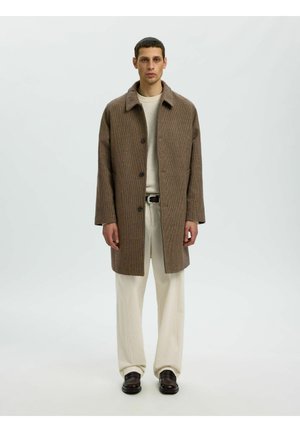 Homme portant un pardessus marron texturé, un pull crème, un pantalon crème à jambes larges, une ceinture noire et des chaussures habillées marron foncé, debout devant un fond uni.