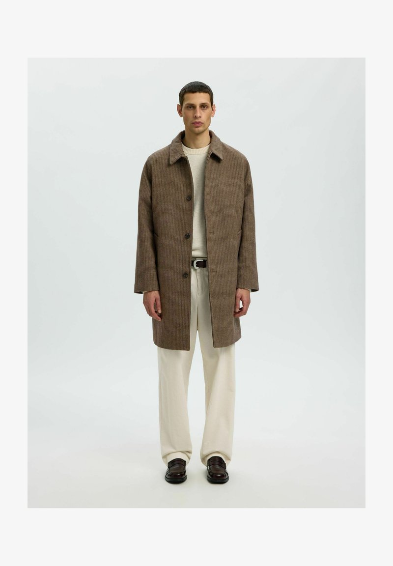 Homme portant un pardessus marron texturé, un pull crème, un pantalon crème à jambes larges, une ceinture noire et des chaussures habillées marron foncé, debout devant un fond uni.