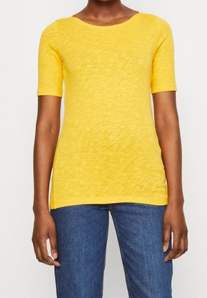 Person iført en ensfarvet gul kortærmet t-shirt og blå jeans, der står mod en hvid baggrund.