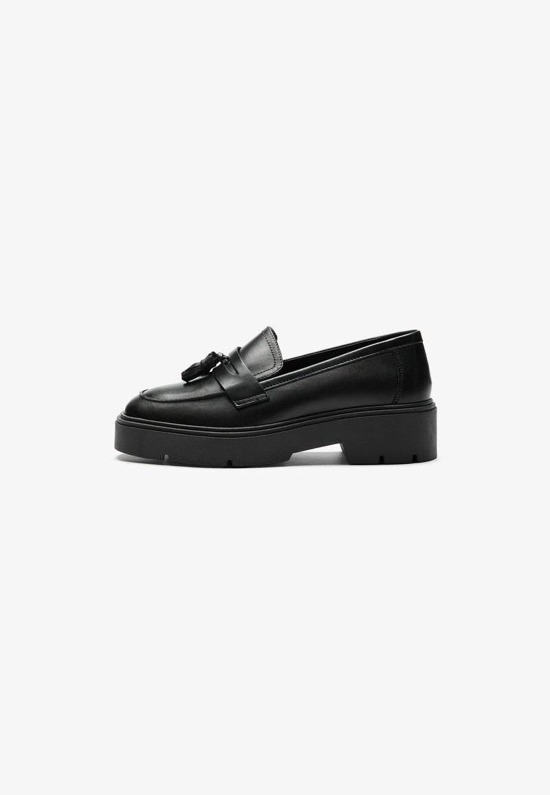 Mocassins de couro preto com uma fivela proeminente e detalhe de franjas, apresentando uma sola plataforma espessa e texturizada. Acabamento suave e perfil baixo.