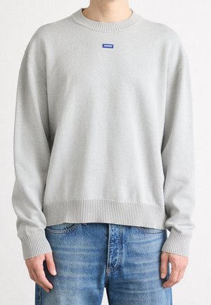 Hellgrauer Pullover mit langen Ärmeln, gerippten Bündchen und Saum, kleines blaues "HUGO"-Logo auf der Brust, kombiniert mit blauen Jeans.