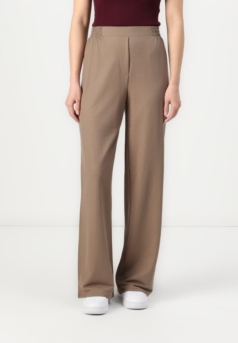 Bruine, wijde broek met een elastische tailleband, gemaakt van een zachte stof, gecombineerd met witte sneakers op een neutrale achtergrond.