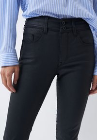 Jeans ajustados negros con acabado brillante, cierre de dos botones y bolsillos estándar. Llevados con una camisa a rayas azul y blanca.