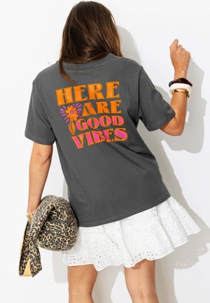 Femme portant un t-shirt gris foncé avec un texte orange et rose "Here are good vibes", tenant une veste à imprimé léopard et portant une jupe blanche à œillets.
