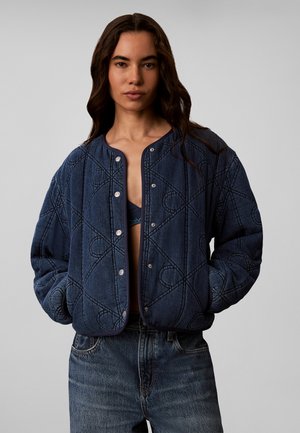 Kvinde iført en mørkeblå quiltet jakke med geometrisk syning, hænderne i lommerne og blå jeans, står mod en ensfarvet baggrund.