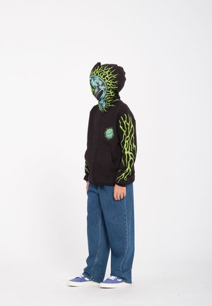 Zwarte hoodie met ritssluiting, voorzien van groene vlamachtige accenten en een afbeelding op de voorkant. Gecombineerd met losse blauwe jeans en paarse sneakers.