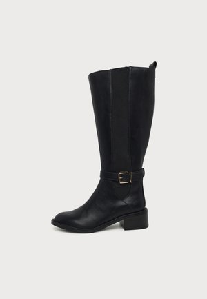 Botte noire montant jusqu’au genou, avec un empiècement latéral élastique, une bride à la cheville et une boucle dorée, sur un fond clair uni.