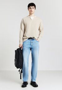 Camicia beige a maniche lunghe con scollo a V e tre bottoni, abbinata a jeans azzurri a gamba dritta. Zaino nero e scarpe nere lucide.