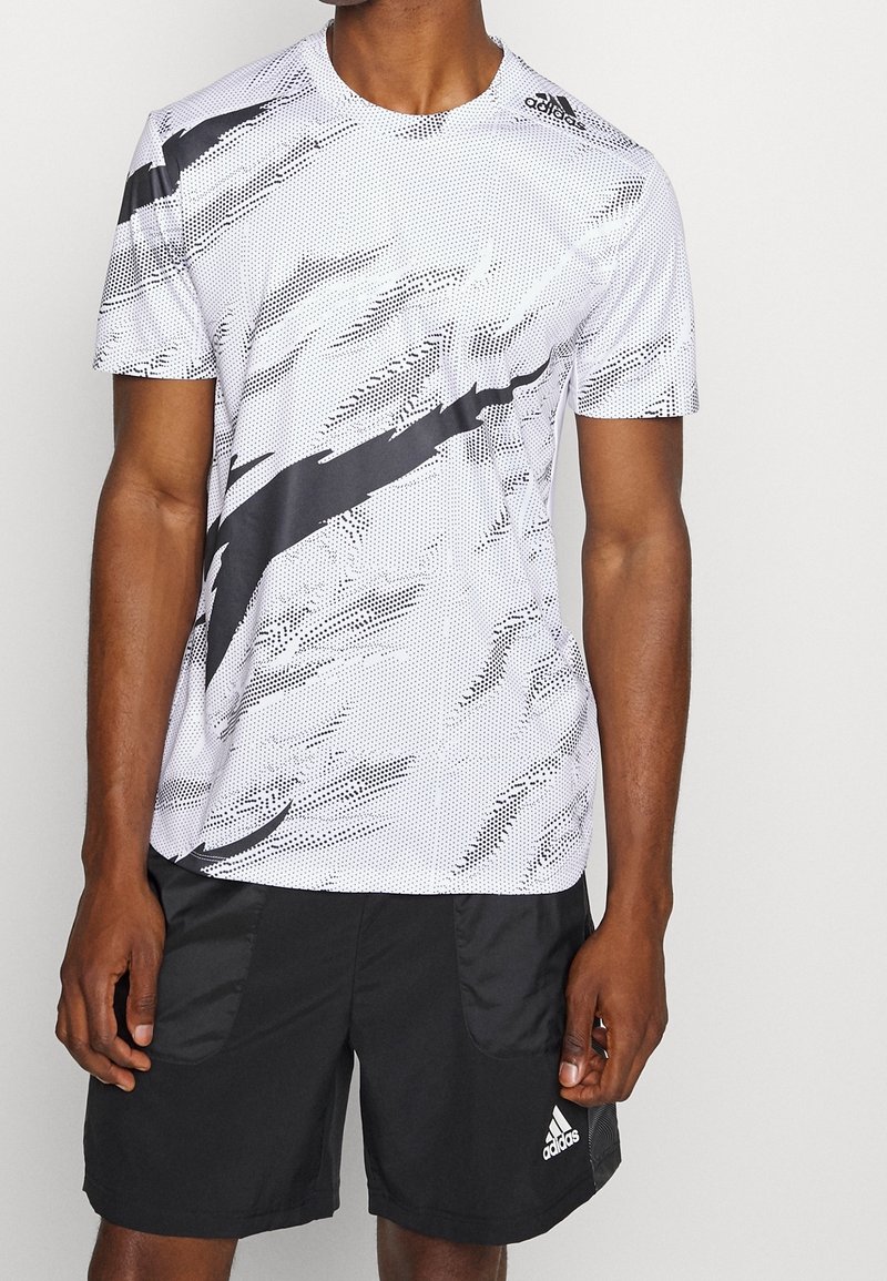 Camiseta deportiva blanca con patrón abstracto negro; de manga corta y cuello redondo. Combinada con pantalones cortos negros que tienen bolsillos laterales y un logo.