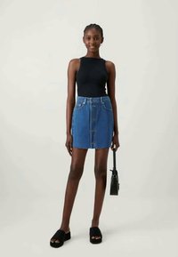 Fekete ujjatlan felső, kék farmer mini szoknya elöl gombokkal, lapos fekete papucs, kis fekete táska a kezében, semleges háttér.