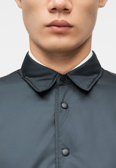 Κοντινό πλάνο του κάτω μέρους του προσώπου και του λαιμού ενός ατόμου που φοράει σκούρο πουκάμισο με κουμπιά και μυτερό γιακά πάνω από λευκό εσώρουχο.