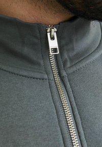 Jachetă gri cu fermoar, având un guler înalt și un fermoar argintiu. Materialul are o textură netedă, cu detalii de cusătură vizibile.