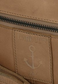 Etichetta in pelle marrone con il design a rilievo di "HARBOUR" e di un'ancora, con una finitura testurizzata e una cerniera sopra. I bordi cuciti sono visibili.