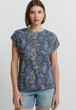 Nuori nainen, jolla on lyhyet ruskeat hiukset, kullanväriset renkaankorvakorut, sininen paisley-kuvioinen lyhythihainen paita ja vaaleansiniset farkkushortsit.