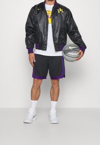 Chaqueta bomber de satén negra con texto amarillo "LAL" y acentos morados, combinada con pantalones cortos grises con ribetes morados. Sosteniendo un balón de baloncesto gris de Nike.