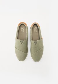 Zapatos verdes tipo slip-on con una parte superior de tela texturizada, que presentan un frente en forma de V y detalles en color marrón claro en el talón, y una suela de goma blanca.