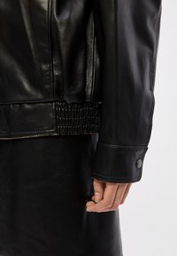 Crna kožna jakna s nabranim pojasom, glatkom teksturom, širokim manžetama s gumbima na preklop te krupnim planom ruke.