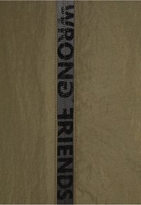 Olivgrüner Stoff mit strukturierten Oberflächen, der ein schwarzes Band in der Mitte aufweist, auf dem die Worte "FALSCHE FREUNDE" in fetter Schrift stehen.