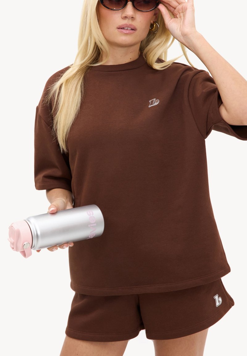 Femme blonde portant un short marron et un T-shirt, tenant une bouteille d’eau argentée avec un bouchon rose, ajustant ses lunettes de soleil, fond clair uni.