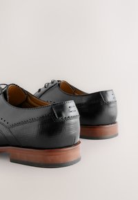 Black leather dress shoes feature a smooth finish, decorative stitching, and a brown wooden heel with a contrasting black rubber sole.