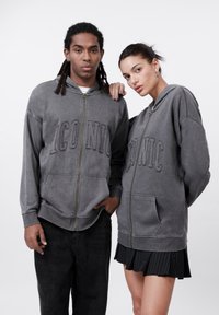 Siva zip-up dukserica s natpisom "IKONIČNO" na prednjoj strani. Ima kapuljaču, prednje džepove i opušteni kroj. Model nosi crne traperice i plisiranu suknju.