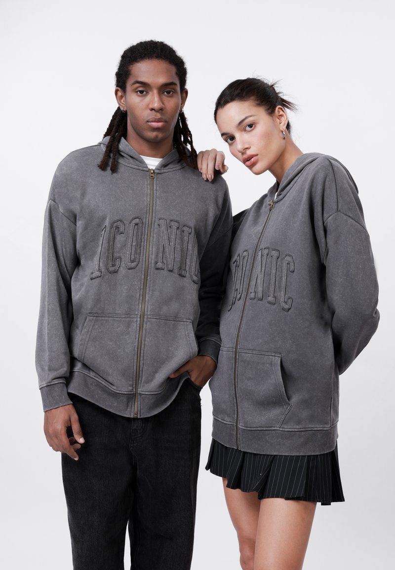 Siva zip-up dukserica s natpisom "IKONIČNO" na prednjoj strani. Ima kapuljaču, prednje džepove i opušteni kroj. Model nosi crne traperice i plisiranu suknju.