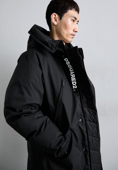 Schwarze Daunenjacke mit hohem Kragen, Reißverschluss-Tasche und weißem Logo auf der Vorderseite. Das Material wirkt glatt und synthetisch.