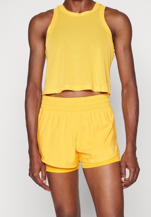 Atletisch persoon draagt een mouwloze gele mesh crop top en bijpassende gele hardloopshorts met elastische tailleband.