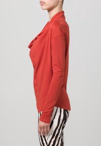 Koszula z długim rękawem w kolorze rdzy, wykonana z bawełny, z drapowanym przodem. Noszona z spodniami w czarno-białe paski. Gładka tekstura, luźny krój.