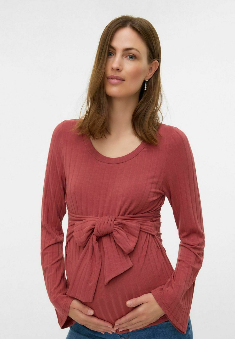 Top gravidanza a maniche lunghe in tessuto a costine di colore rosso ruggine, con dettaglio nodo in vita, abbinato a jeans blu. Tessuto morbido e vestibilità comoda.