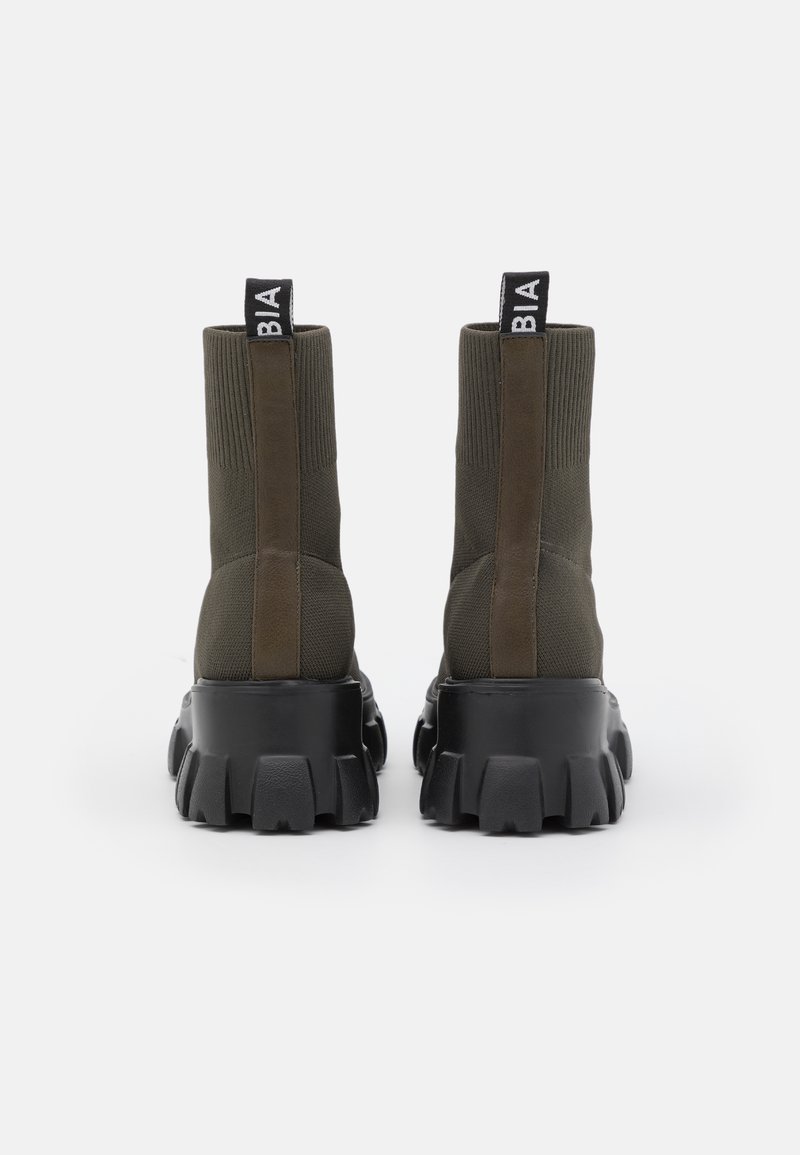 Une paire de bottes-socquettes vert olive mi-mollet avec des semelles en caoutchouc noires épaisses, une bande de cuir verticale et des languettes en tissu avec du texte blanc à l'arrière.
