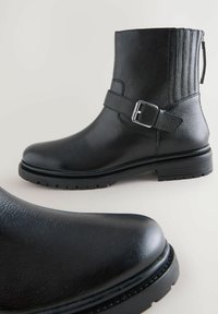 Botín negro de cuero hasta el tobillo con acabado texturizado, cremallera lateral, correa decorativa con hebilla y suela de goma robusta.