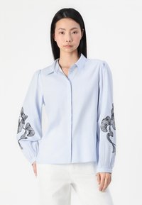 Camisa azul-clara com botões, com bordado floral preto em ambas as mangas. Apresenta colarinho, mangas longas e bufantes, e um corte descontraído.