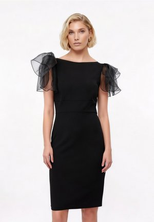 Femeie cu păr blond scurt poartă o rochie neagră mulată cu mâneci bufante translucide în straturi și cercei aurii în formă de cerc, stând în fața unui fundal simplu.