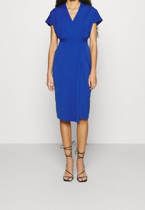 Femme portant une robe portefeuille bleue mi-longue avec des manches courtes et des sandales à talons hauts noires à brides, debout devant un fond uni.