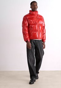Red puffer jacket with a shiny finish, high collar, front zip, and two side pockets. Worn with dark wide-leg jeans and black shoes.