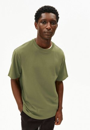 Junger Mann steht mit den Händen in den Taschen, trägt ein olivgrünes T-Shirt mit kurzen Ärmeln und dunkle Hosen vor weißem Hintergrund.