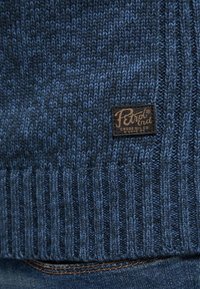 Granatowy sweter dzianinowy z prążkowaną fakturą u dołu oraz prostokątną metką z logo i nazwą marki.