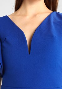 Kungligt blå klädsel med djup V-ringning och långa ärmar, tillverkad av slätt tyg. Har en figurnära silhuett och rena linjer.