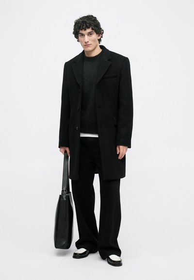 Άνδρας με σγουρά σκοτεινά μαλλιά, φορώντας μαύρο παλτό, μαύρο πουλόβερ, μαύρο παντελόνι, μαύρα παπούτσια με λευκή σόλα, και κρατώντας μια μεγάλη μαύρη δερμάτινη τσάντα.