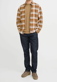 Camisa de franela a cuadros en beige y naranja, superpuesta sobre una camiseta marrón, combinada con unos pantalones vaqueros azul oscuro y zapatos de ante marrón.