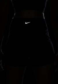 Juodi sportiniai šortai su atspindinčiu baltu Nike logotipu. Medžiaga atrodo lygi, tvirtai priglundanti prie liemens ir šlaunų.