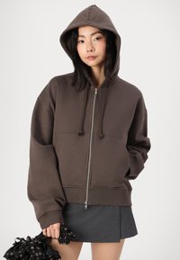 Sudadera marrón con cremallera, gran capucha, mangas largas y bolsillos frontales; combinada con una falda gris oscura. Material de mezcla de algodón texturizado.