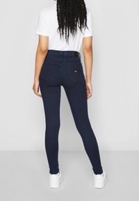 Jeans oscuros ajustados con una cintura alta, que presentan dos bolsillos traseros y un detalle de costura sutil, combinados con una camiseta blanca y zapatillas blancas.